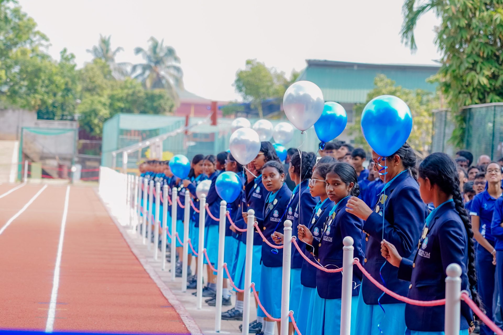 Schoolgirls in blue uniforms holding balloons lined up beside a running track at school event.