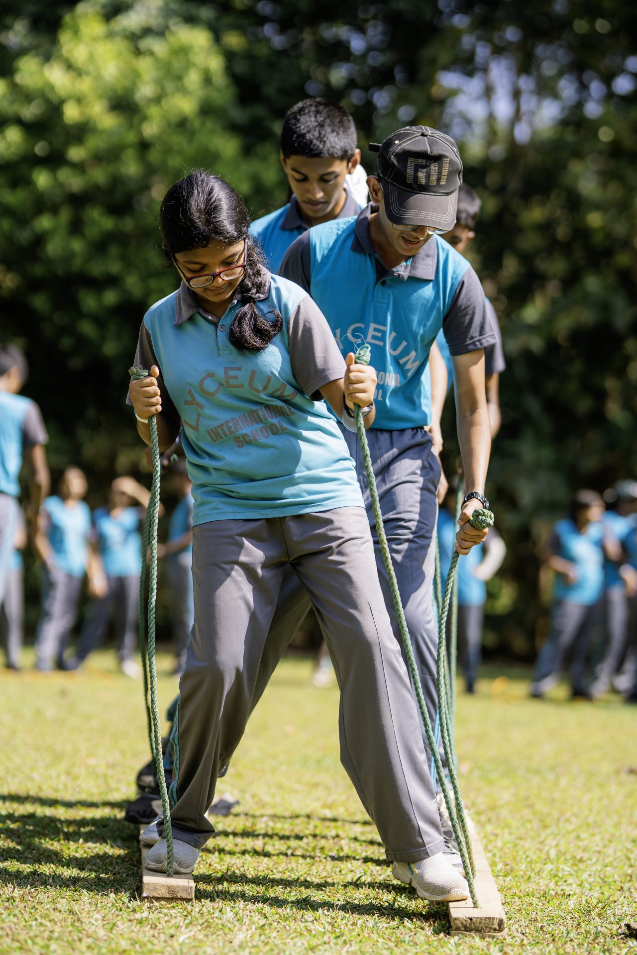 Students in blue shirts balance together on wooden planks during an outdoor team-building activity.