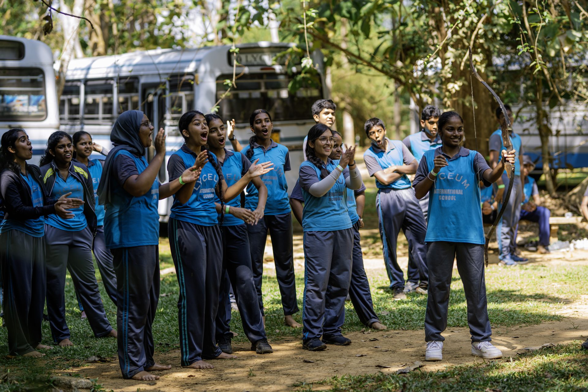 Students in blue shirts cheer and bond outdoors during a lively school field trip under the trees.
