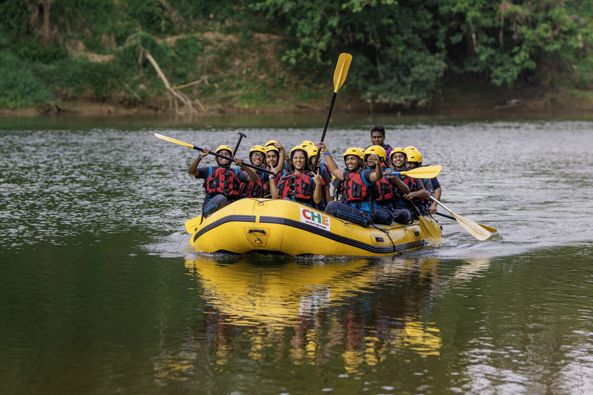 Group rafting on calm river at CHE Adventure Park, wearing helmets and life jackets, smiling.