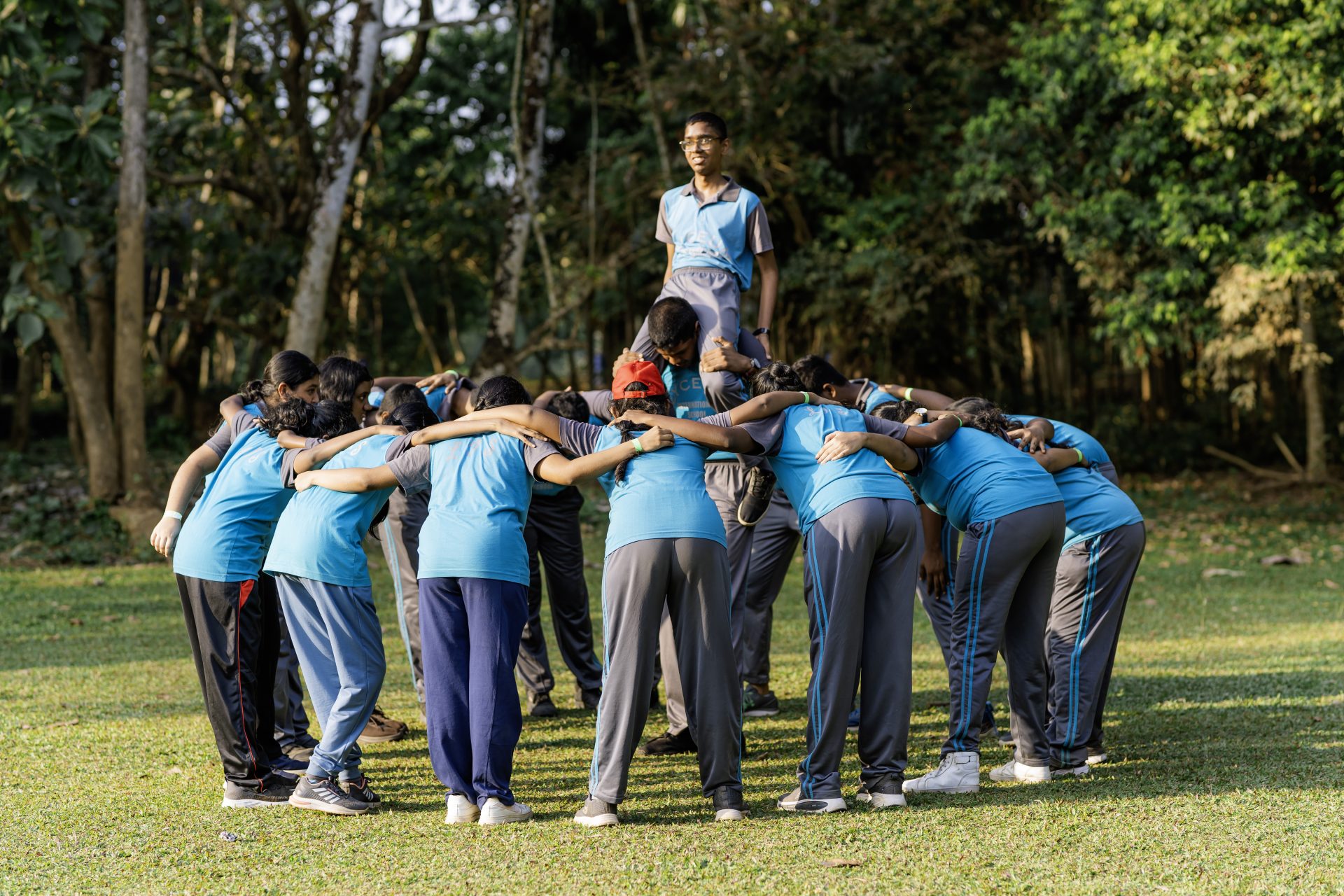 Students in blue sportswear lift teammate during team-building activity on a sunny, grassy field.
