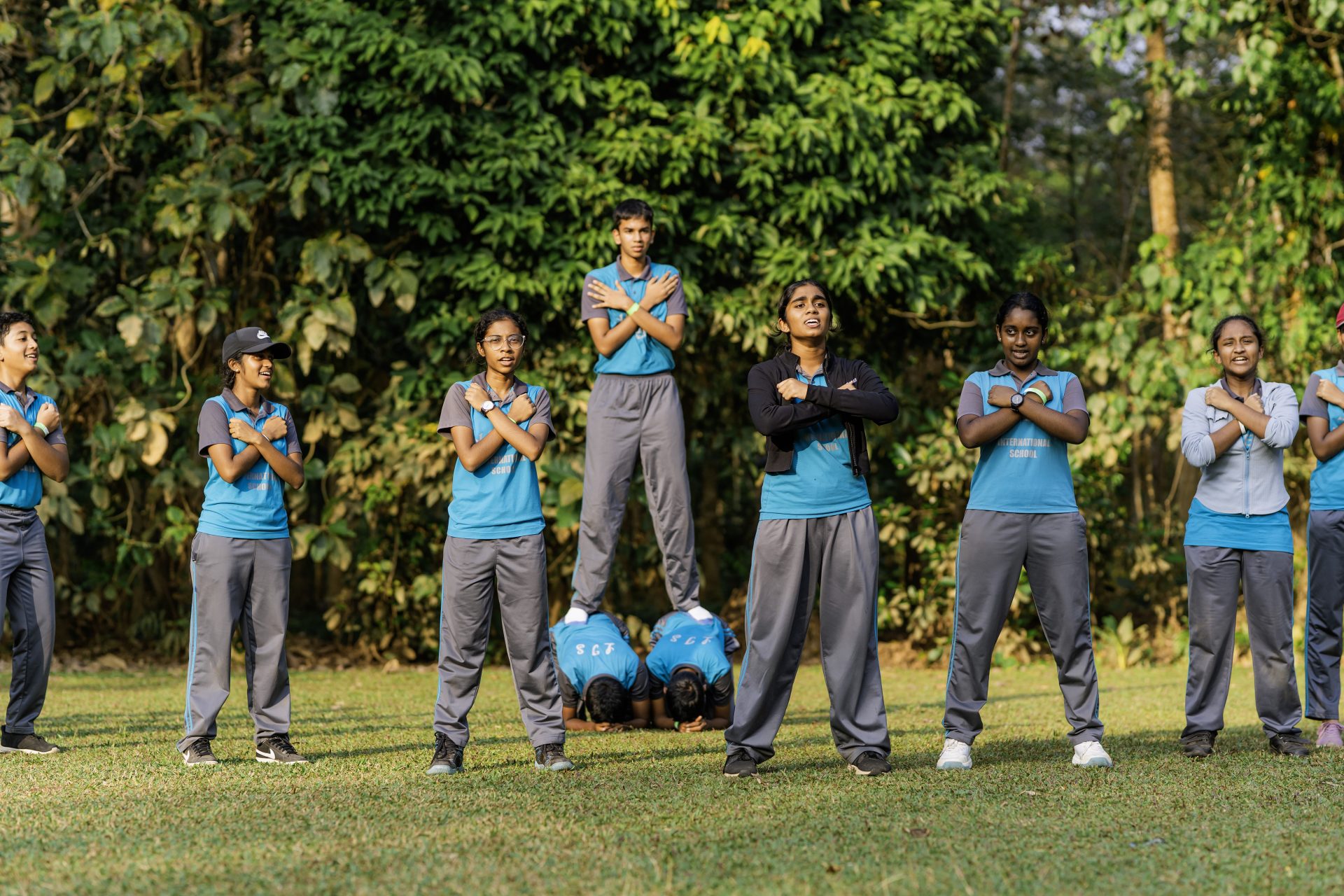 Students in blue uniforms perform a coordinated outdoor balance and teamwork routine in a park.