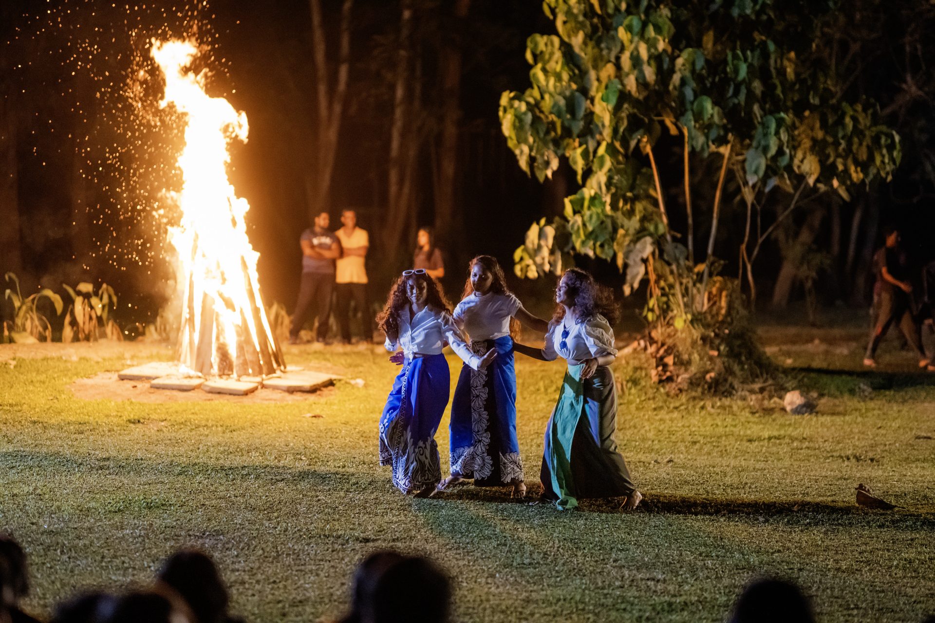 Women in colorful skirts dance by a bonfire at night during a forest cultural festival.