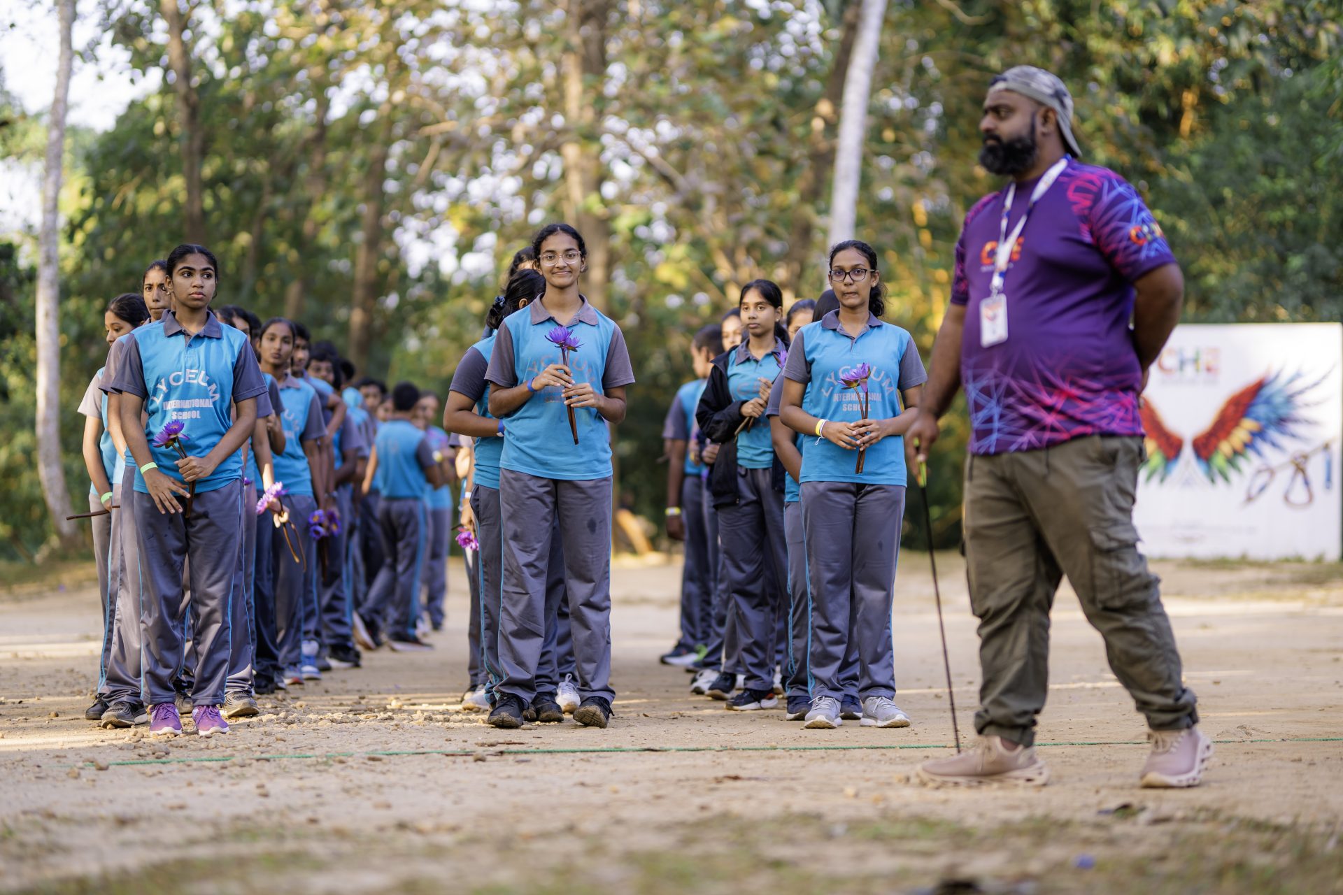 Schoolgirls in blue uniforms hold flowers during outdoor assembly, supervised by teacher in nature.