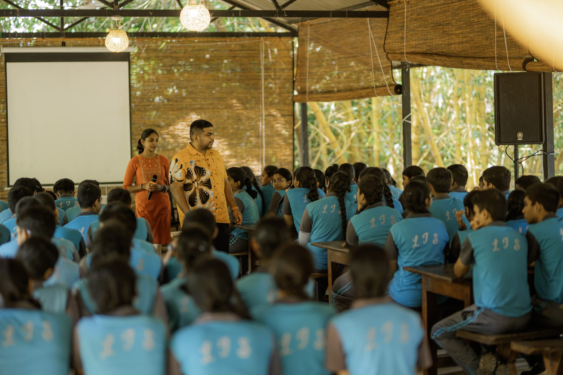 Students in teal uniforms engage in an interactive open-air classroom session with two teachers.