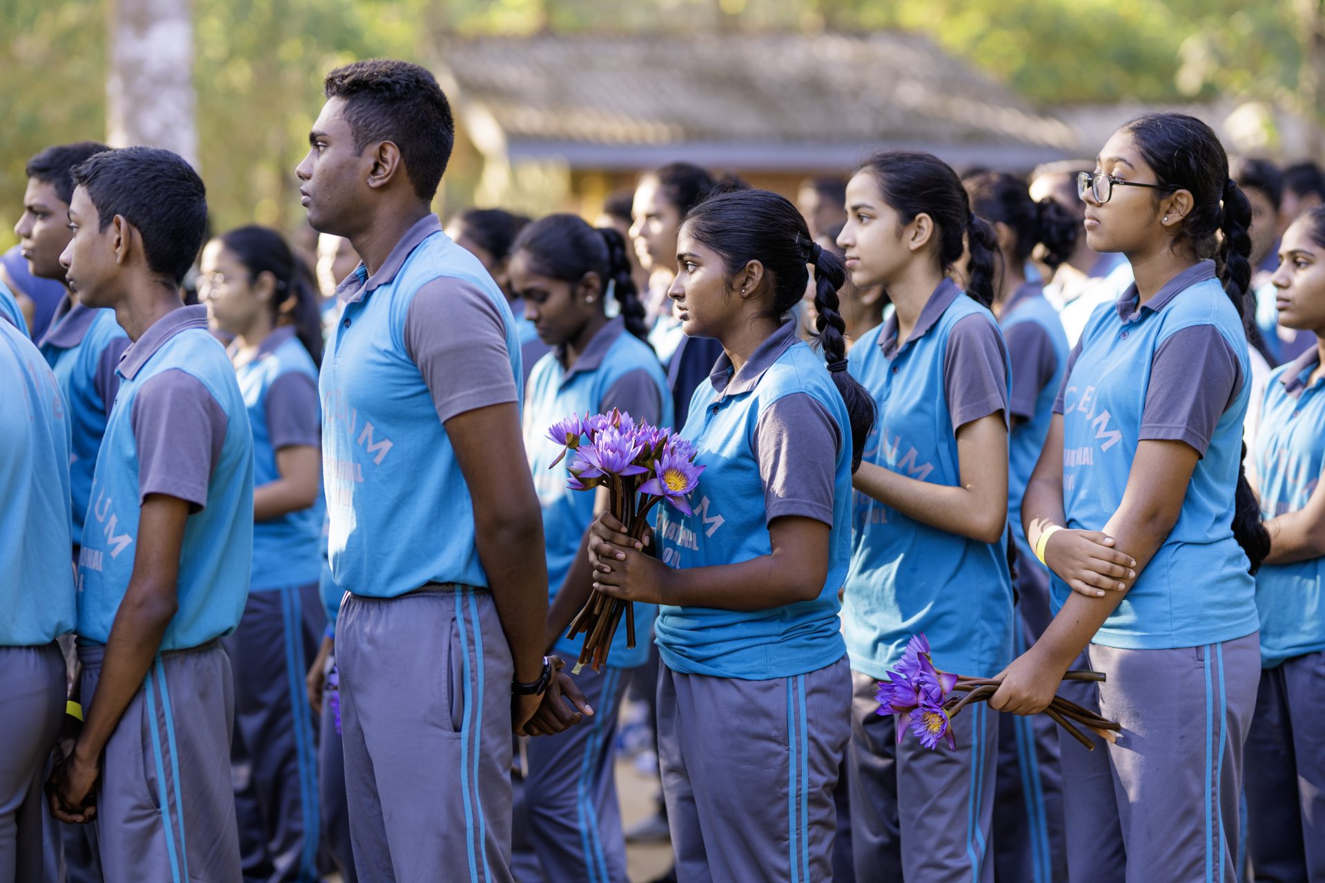 School children in blue uniforms hold purple water lilies during an outdoor ceremony.