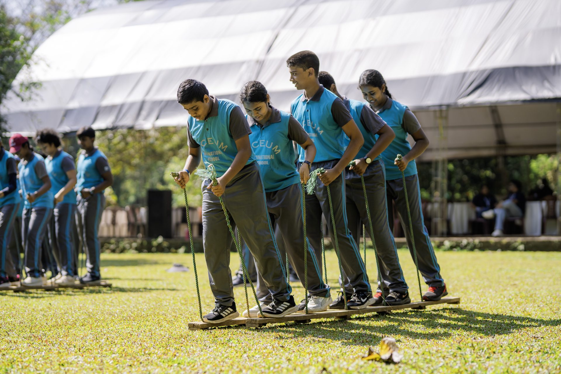 Students in blue vests coordinate on wooden planks during a school outdoor team-building activity.