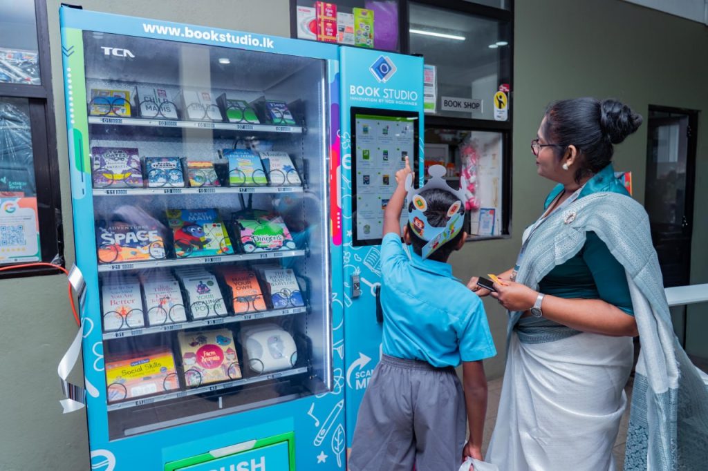 Child picks book from SCHOOL BOOK STUDIO vending machine with teacher, promoting reading and literacy.