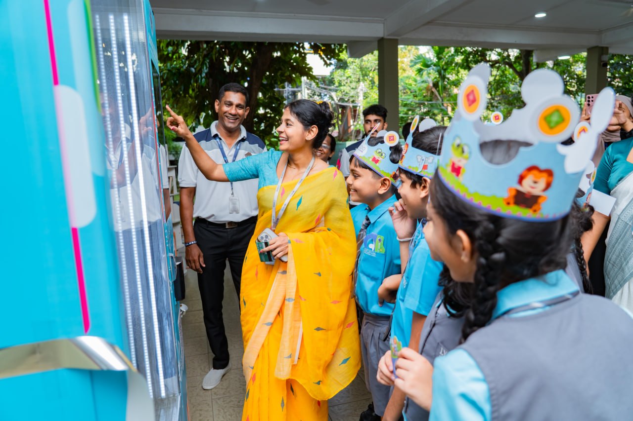 Teacher demonstrates digital kiosk to smiling students in festive crowns at outdoor school event.