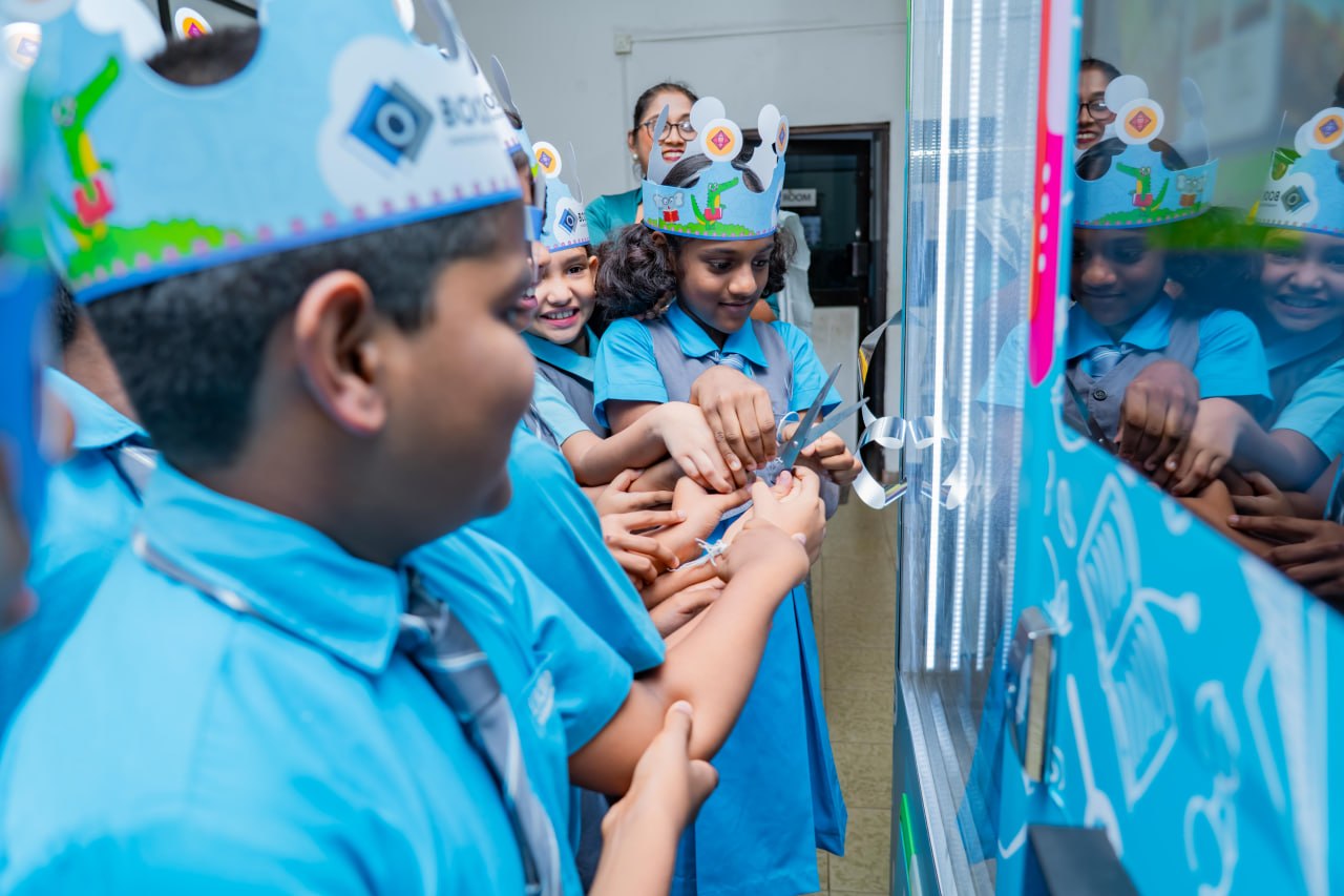 Smiling schoolchildren in blue uniforms cut a ribbon together during a festive classroom celebration.