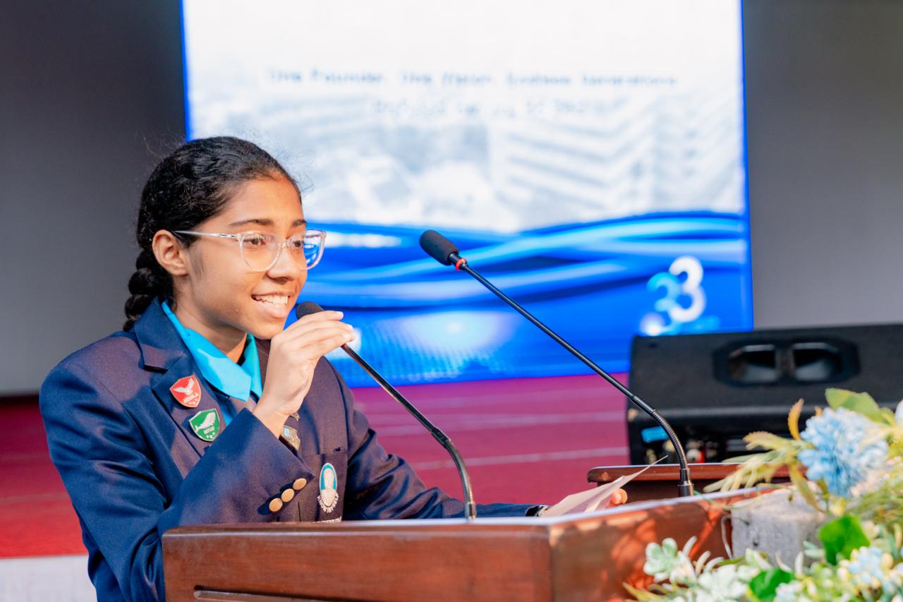 Young girl in navy blazer speaks confidently at podium during academic conference with digital backdrop.