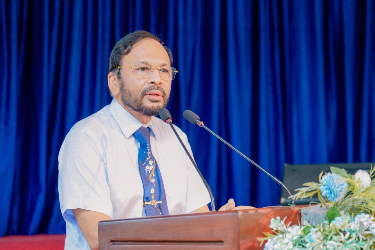 Man in blue shirt and tie speaks at floral podium with blue curtain backdrop at conference.