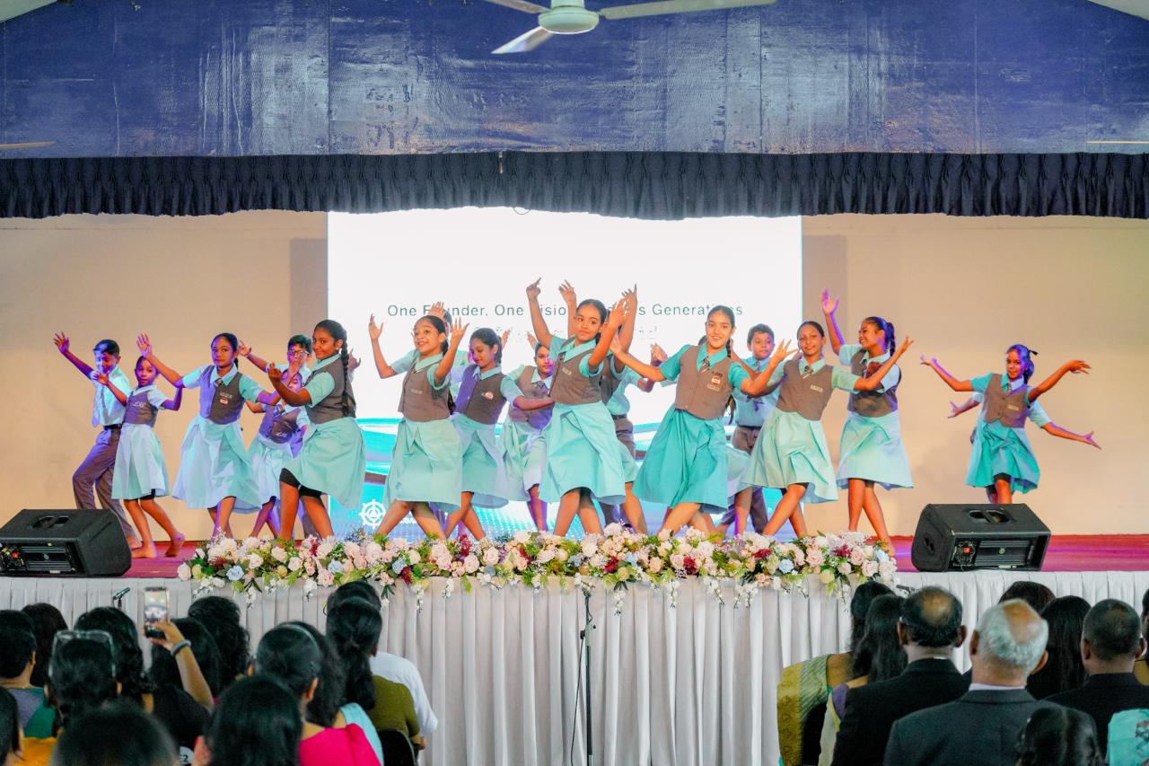 Students in blue costumes perform a lively group dance on stage at a school celebration event.