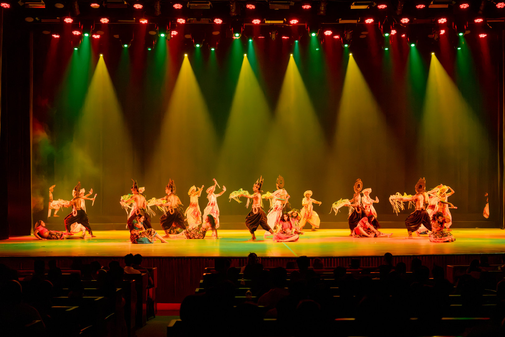 Spectacular Traditional Dance Performance Under Vibrant Stage Lights Dancers in colorful traditional costumes perform on stage with dramatic lights and a visible theater audience.