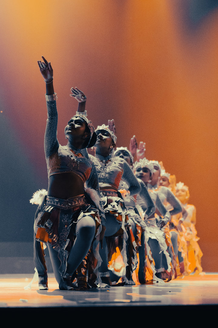 Dynamic Tribal Dance Performance Under Vibrant Stage Lights Synchronized dancers in tribal costumes raise arms on stage under warm, dramatic lighting during cultural performance.