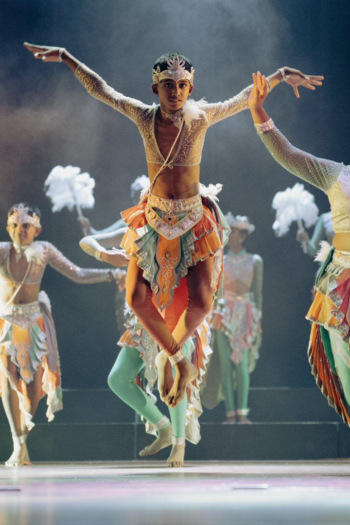 Vibrant Cultural Dance Performance With Colorful Costumes and Leaping Dancer Male dancer in colorful traditional costume leaps on stage with group, dramatic lighting, feather props.