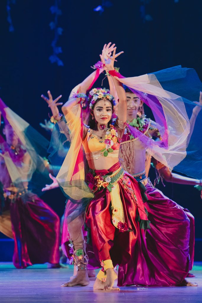 Lead female dancer in vibrant floral costume performs traditional dance with flowing scarves on stage.
