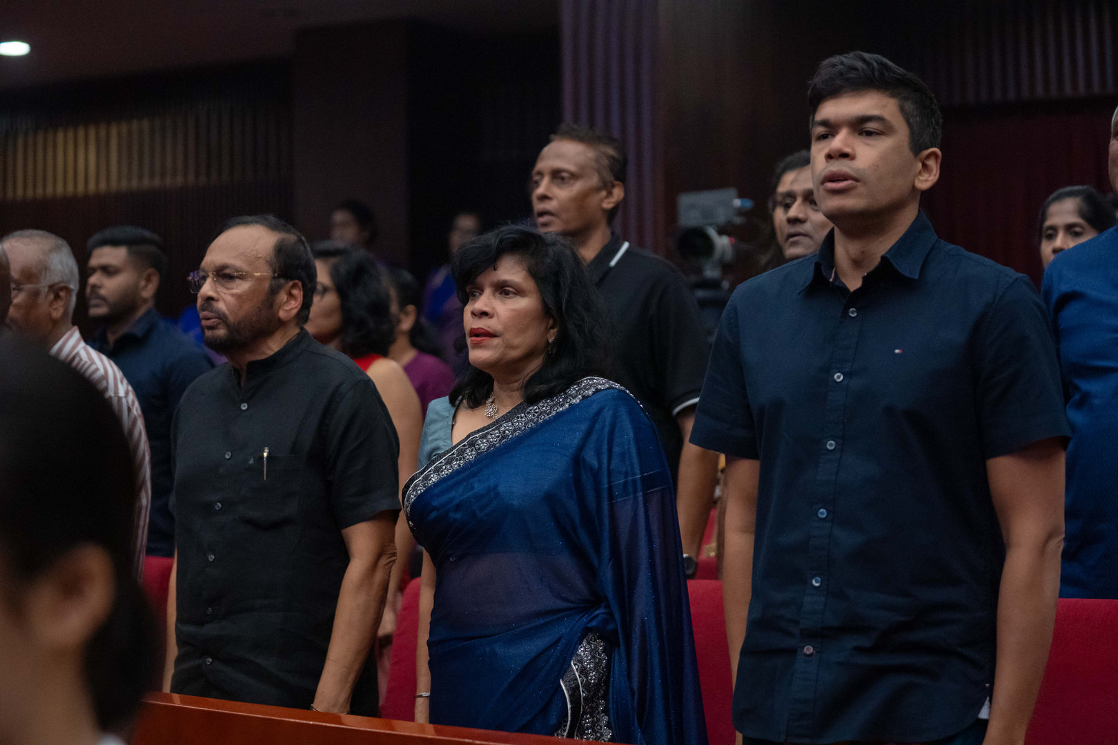 Solemn Ceremony Attendees Standing in Unity and Respect Diverse attendees in formal attire stand respectfully during a solemn ceremony in an auditorium.