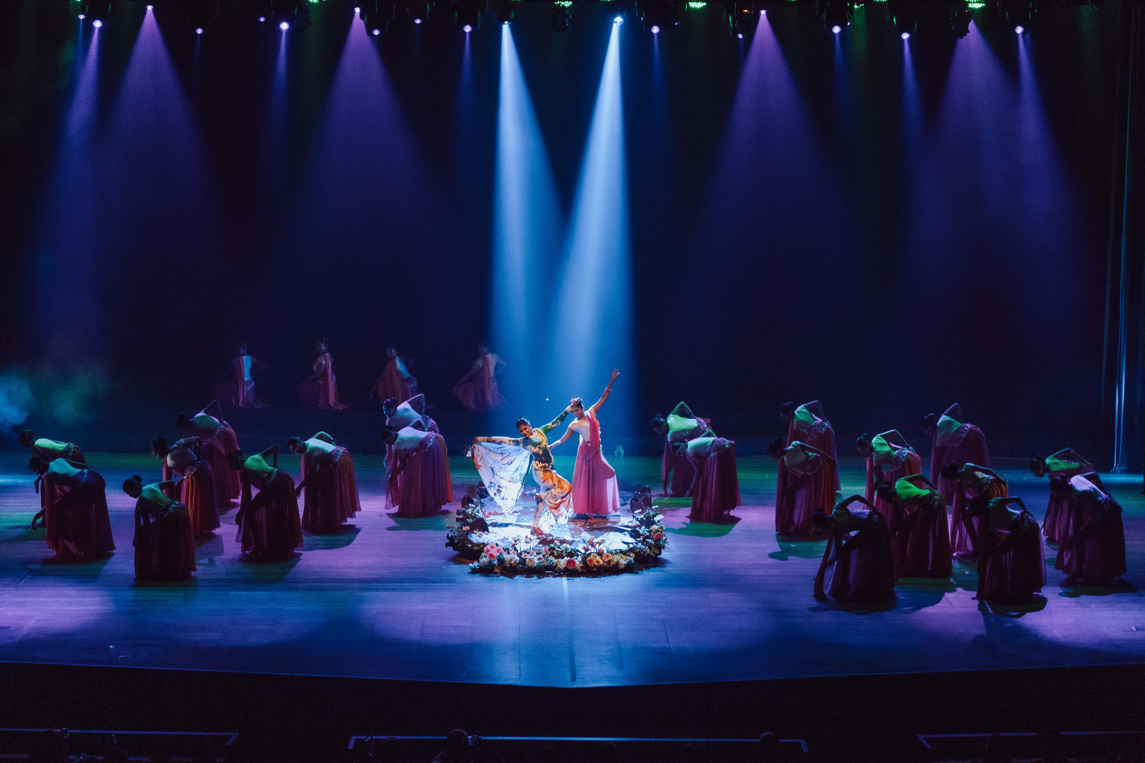 Mystical Stage Dance With Floral Circle and Vibrant Lights Vibrant stage dance with floral platform, dramatic lighting, and performers in colorful traditional costumes.