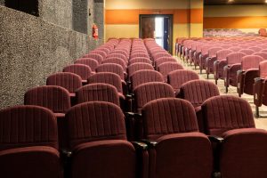 Empty Theater Auditorium With Burgundy Seats and Sunlit Exit Empty theater auditorium with rows of burgundy seats, fire extinguisher, and sunlight through exit doors.